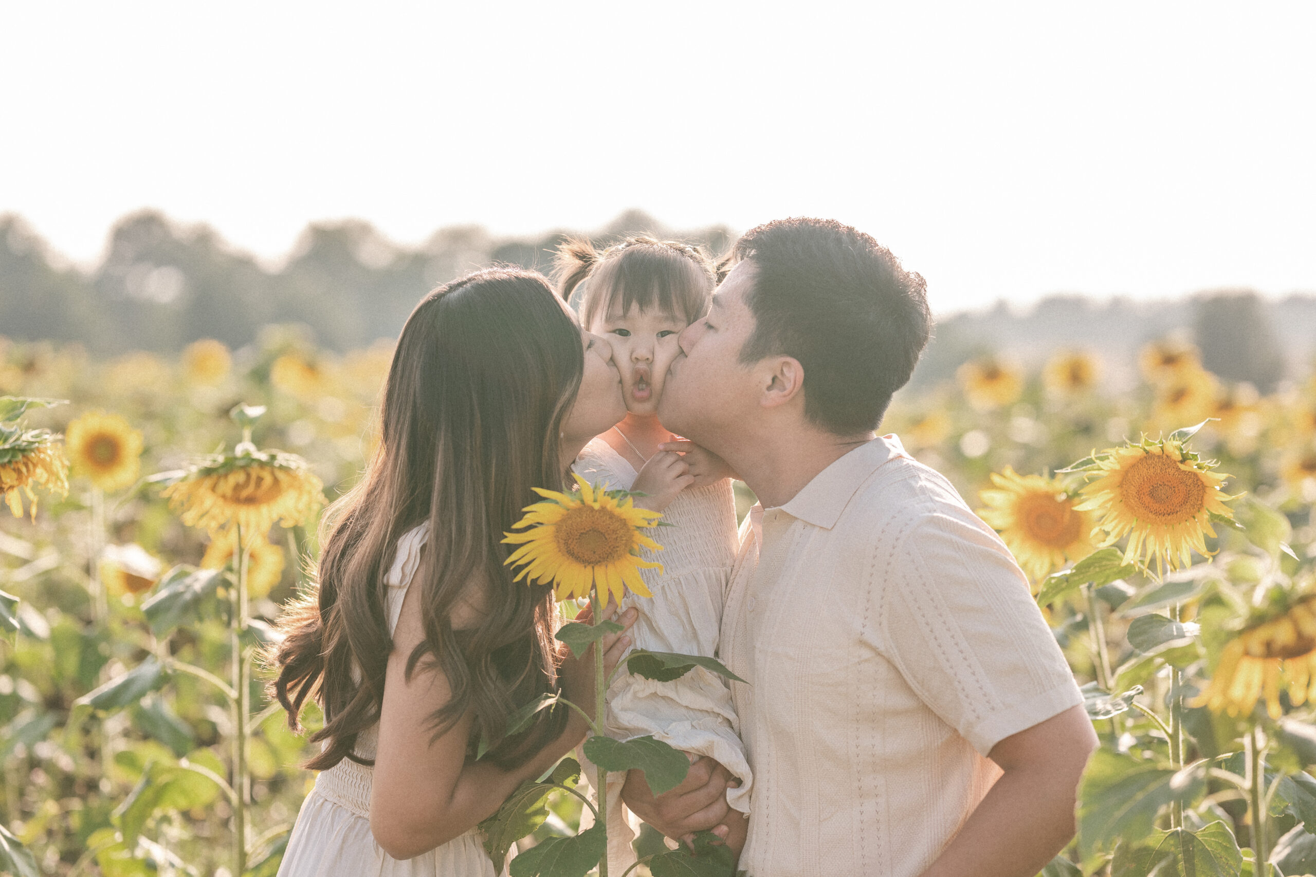 Mom and dad kissing their daughter’s cheeks during seasonal family sessions among the sunflowers.