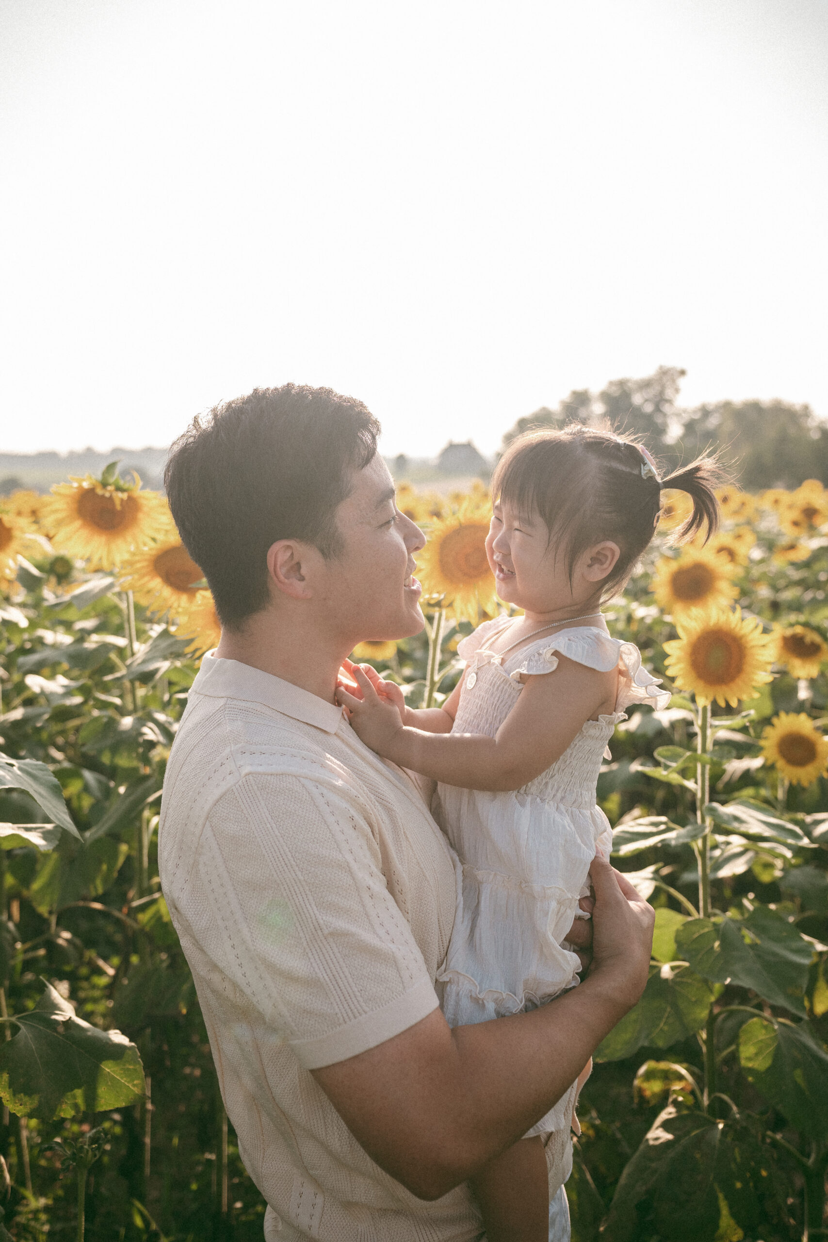 Dad holding his daughter and smiling at her while standing together among rows of sunflowers.