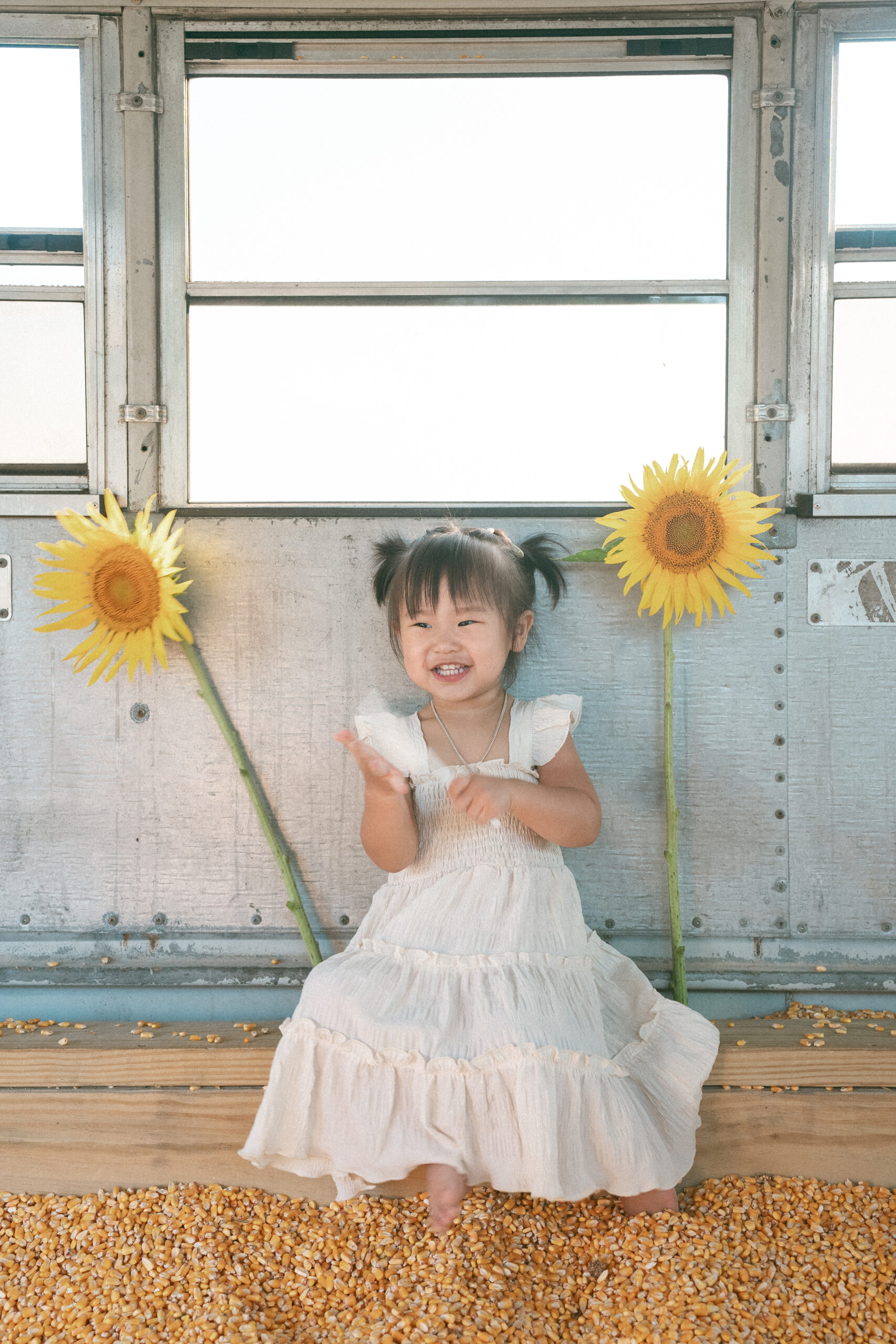 Smiling young girl sitting between two tall sunflowers during seasonal family sessions at a sunflower farm.