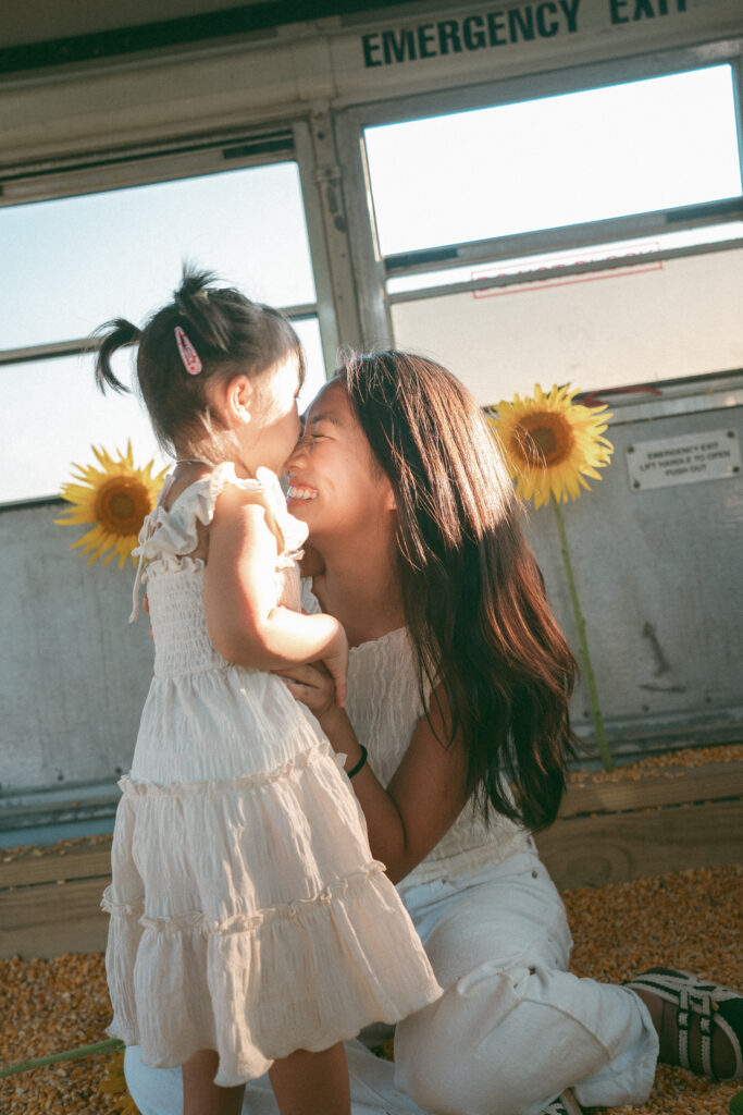 Toddler kisses her mothers forehead with sunflowers in the background.
