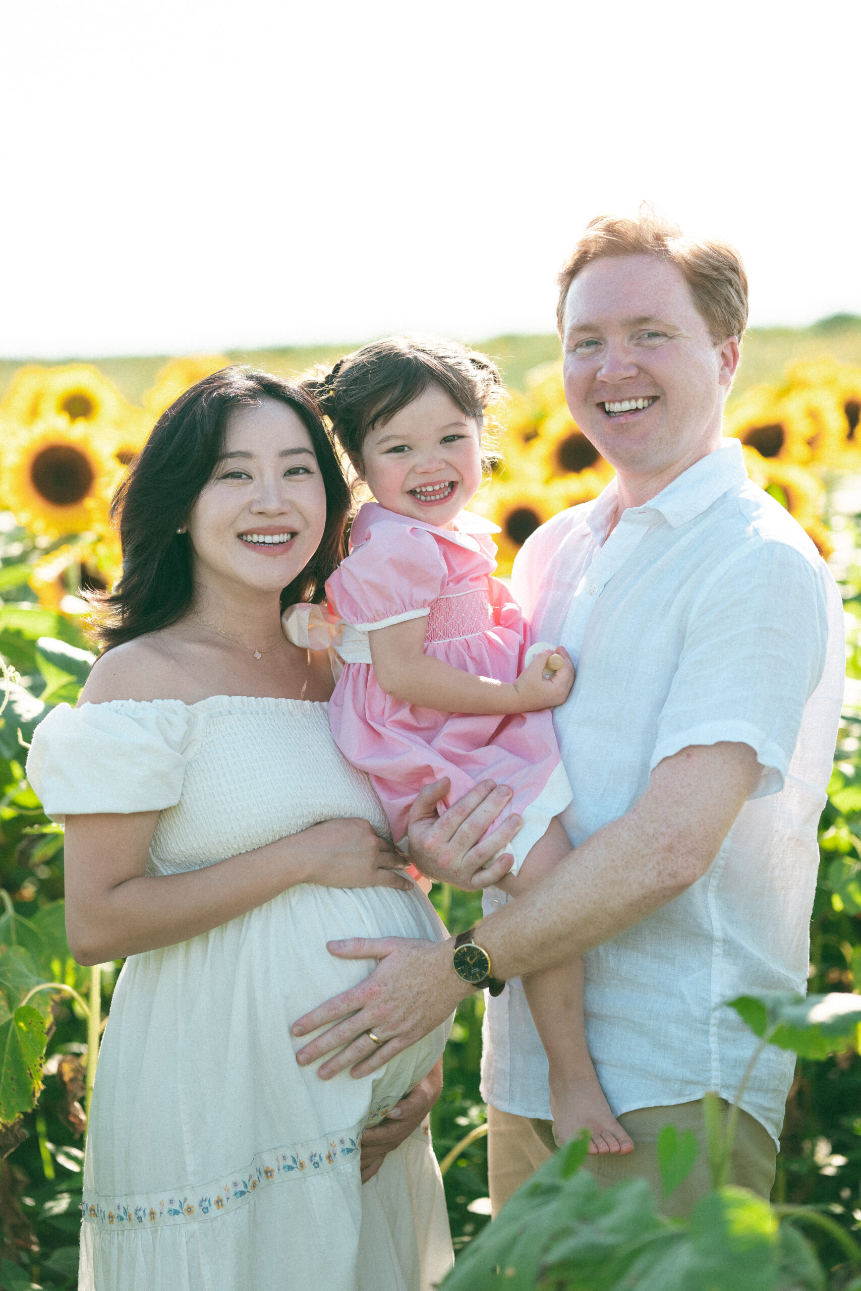 Family of three smiling together in a sunflower field with their young daughter and expecting mother.