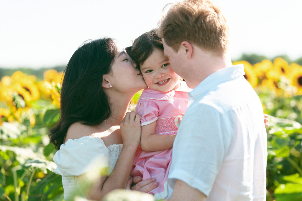 A toddler in the middle of her parents as they kiss her cheek in sunflower fields during their seasonal family sessions.