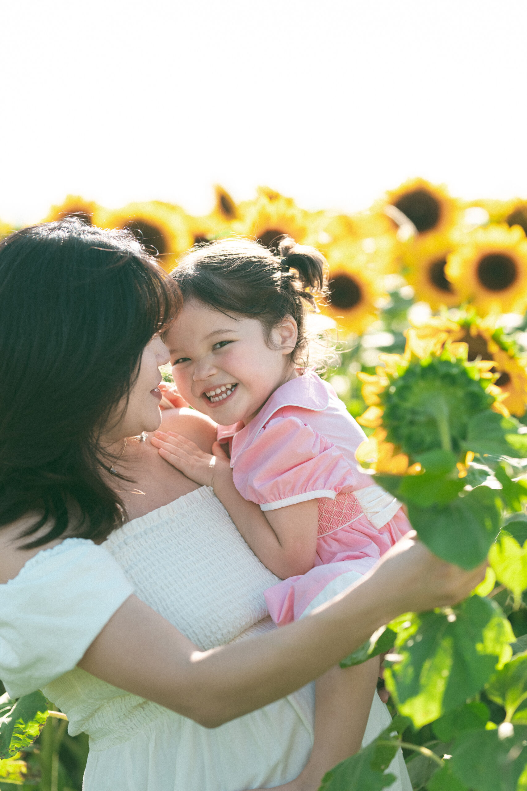 Young girl laughing in her mom’s arms during seasonal family sessions in a sun-filled sunflower field.