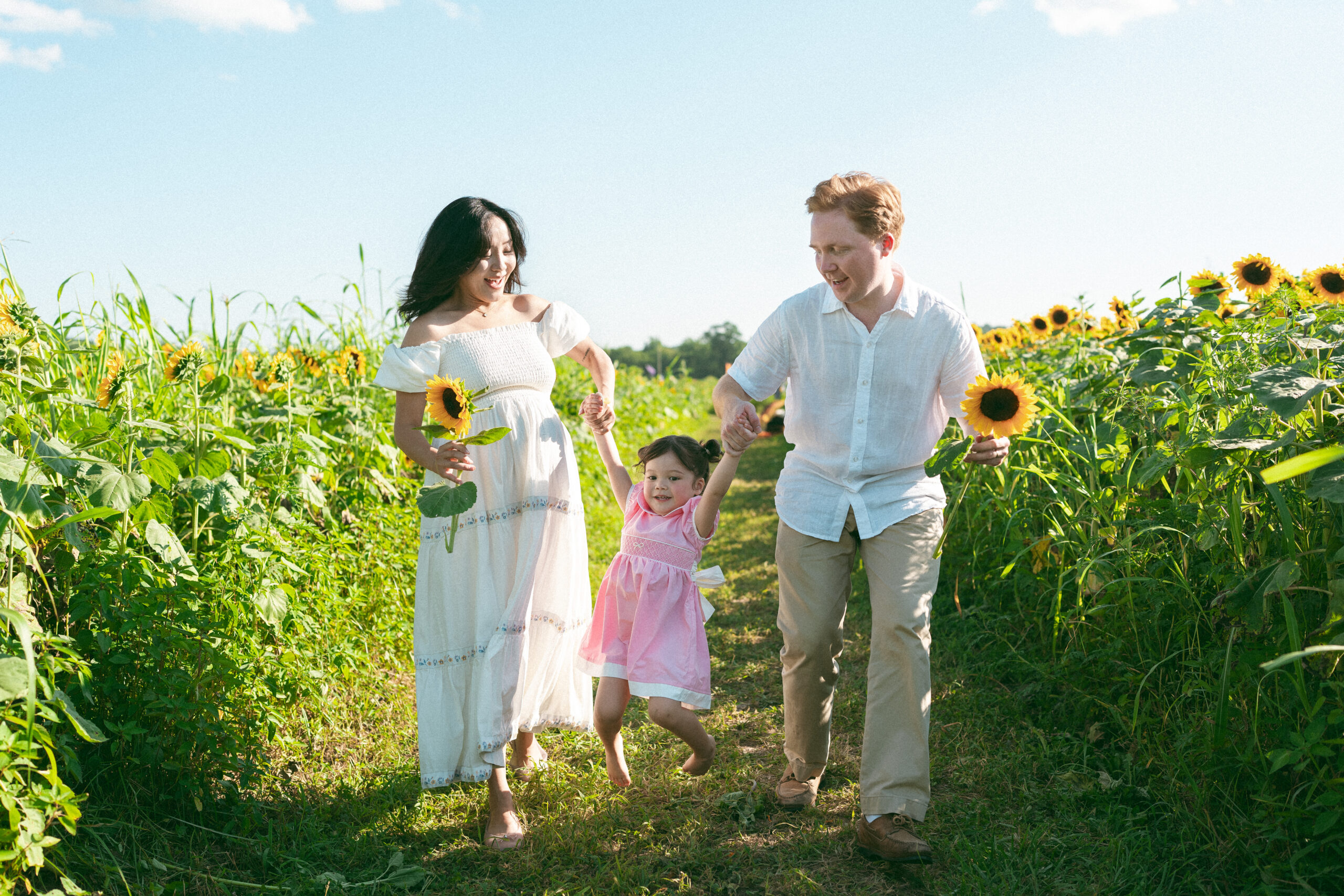 Mom and dad holding their daughter’s hands as they walk together through a sunny sunflower field.
