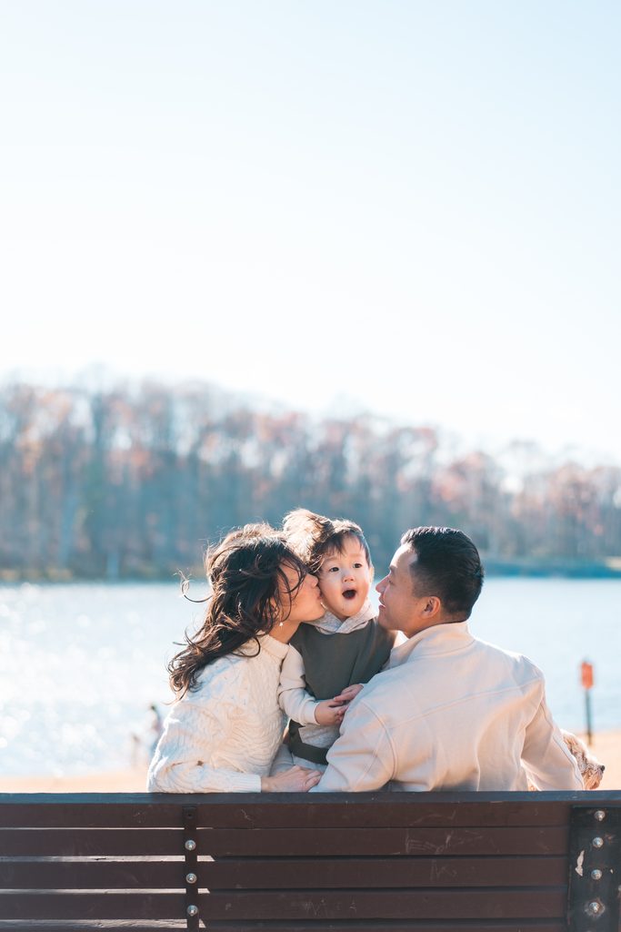 Mom and dad kissing their young son on the cheeks during a sweet family moment by the water as they sit on a bench.