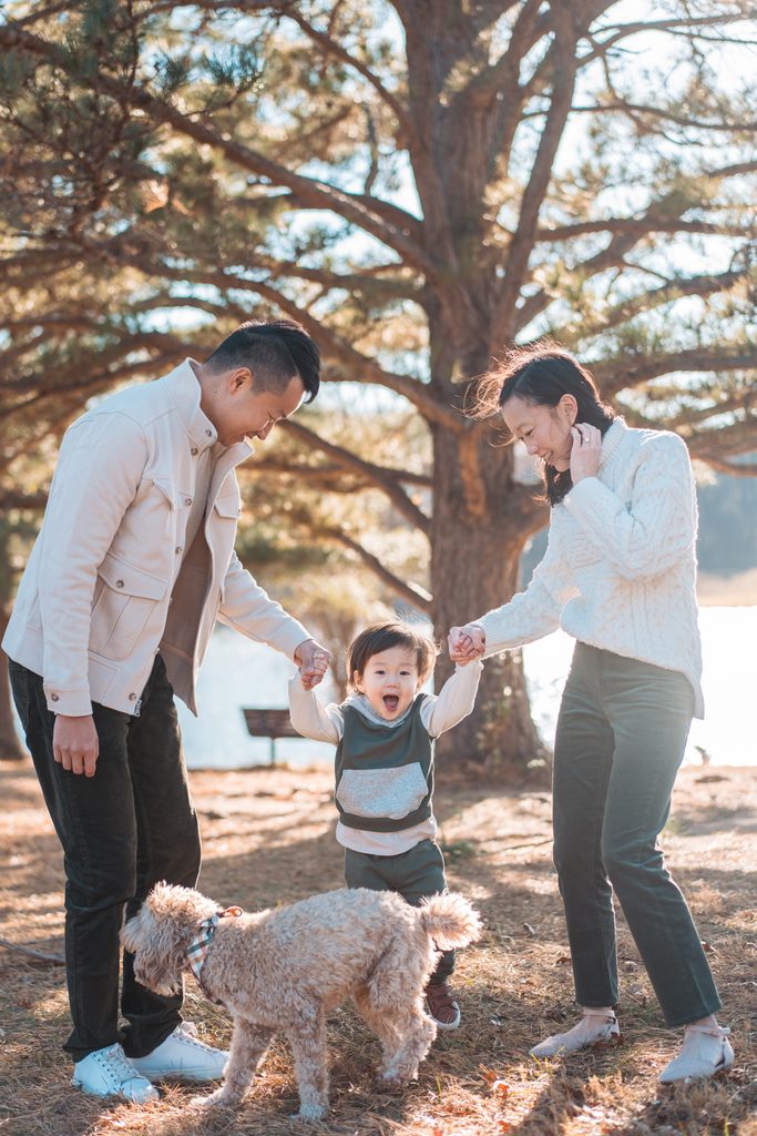 Parents holding their laughing toddler's hands with their dog nearby during Seneca Creek State Park family photos.