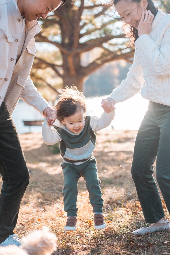 Laughing toddler lifted off the ground while holding both parents' hands during a playful family moment.