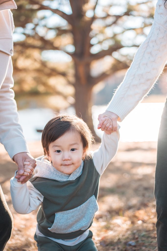 Toddler holding both parents’ hands while walking through the woods during Seneca Creek State Park family photos.