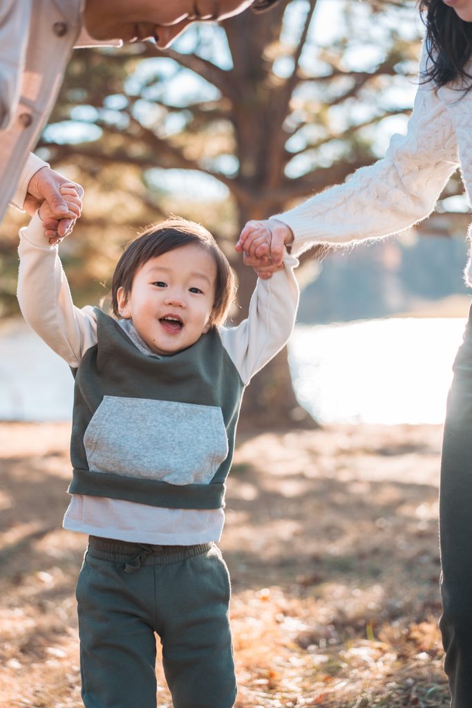 Smiling toddler holding both parents' hands while playing outside near the lake.