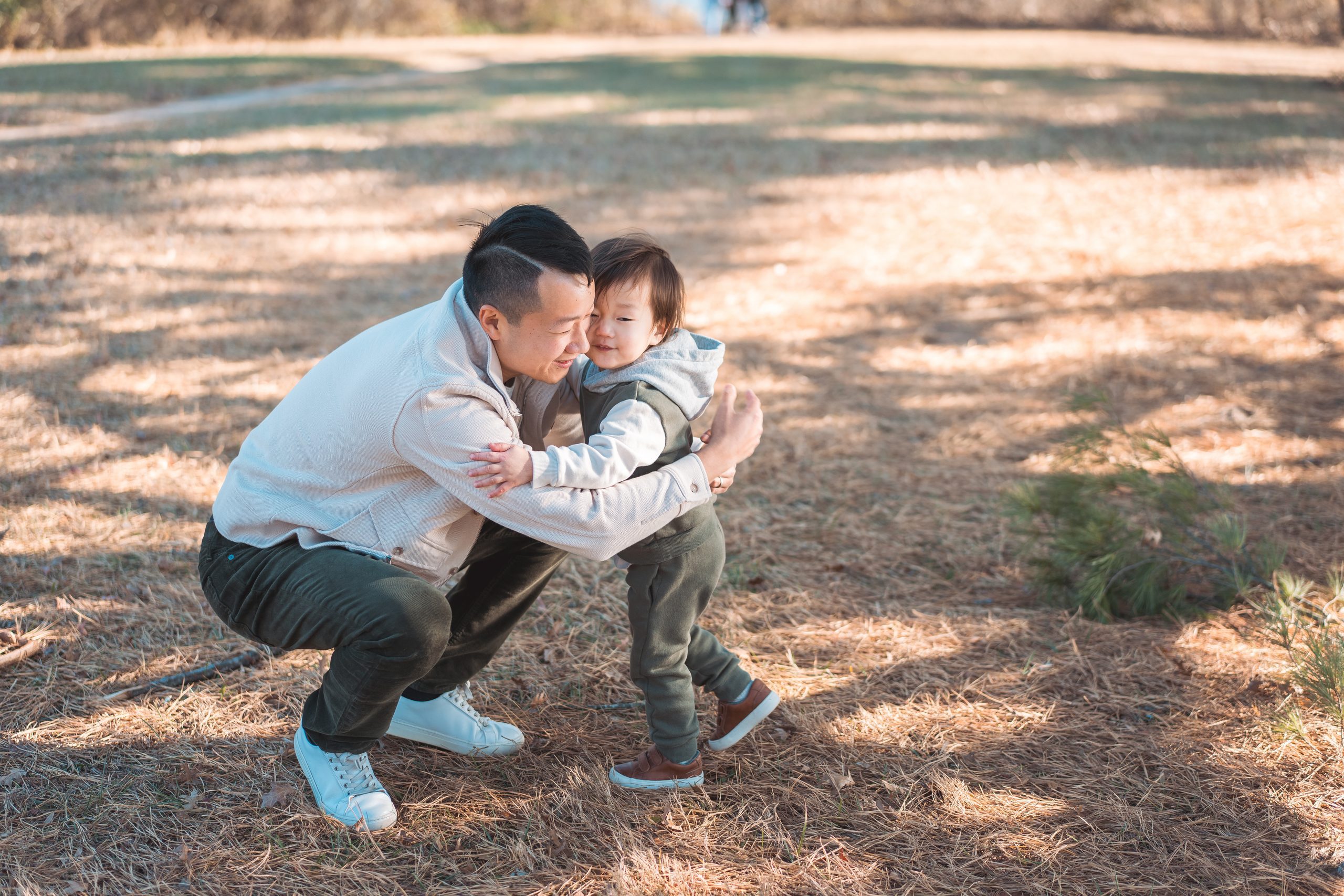 Dad crouching down to hug his young son during candid Seneca Creek State Park family photos in the woods.