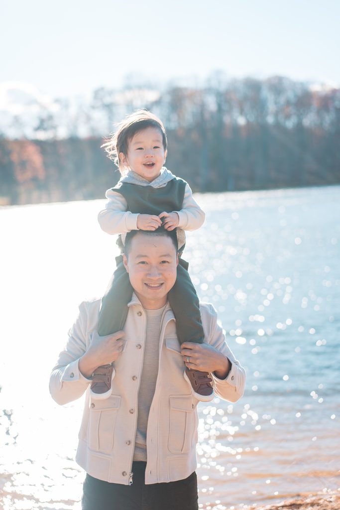Dad carrying his smiling toddler on his shoulders with the sunlit lake behind them.