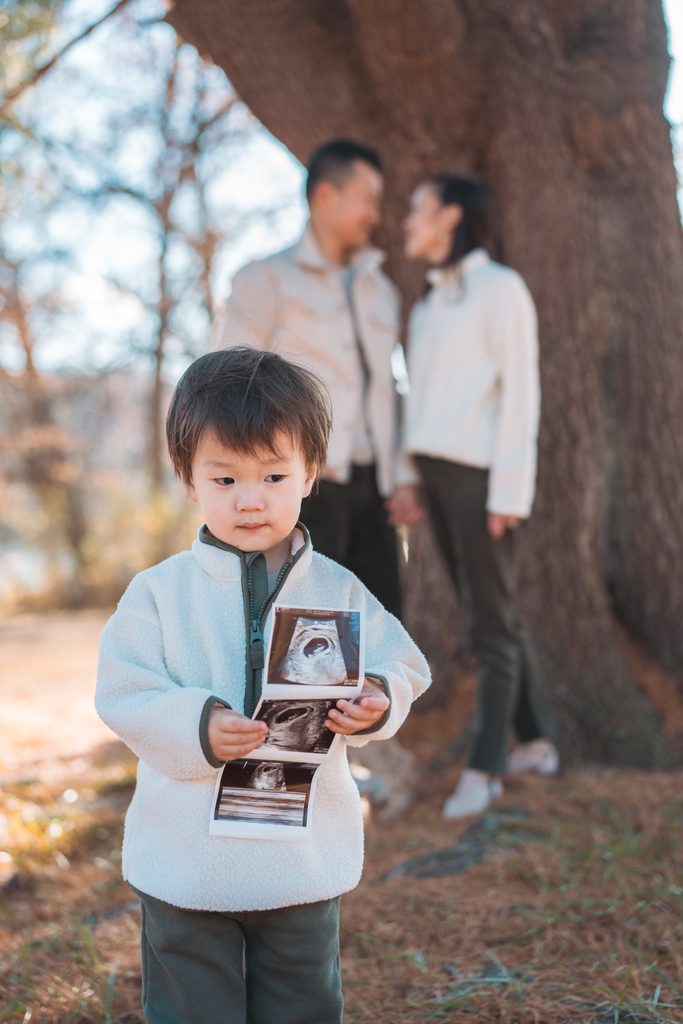 Young boy holding ultrasound photos while his parents share a sweet moment together in the background.