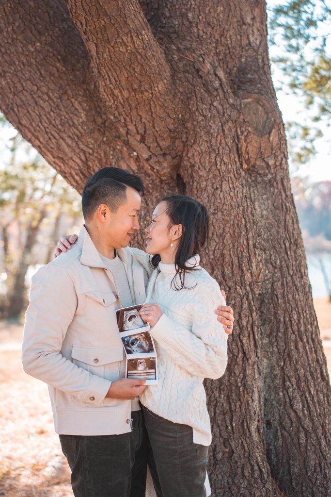 Expecting parents standing together beneath a large tree while holding ultrasound photos during a maternity family session.