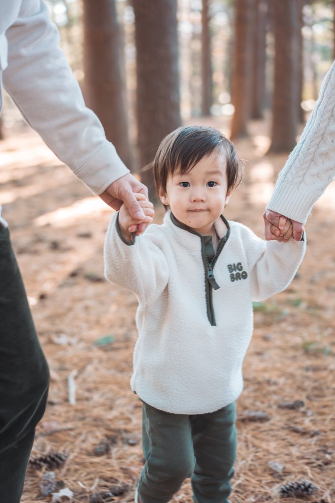 Toddler holding both parents’ hands while walking through the woods during Seneca Creek State Park family photos.