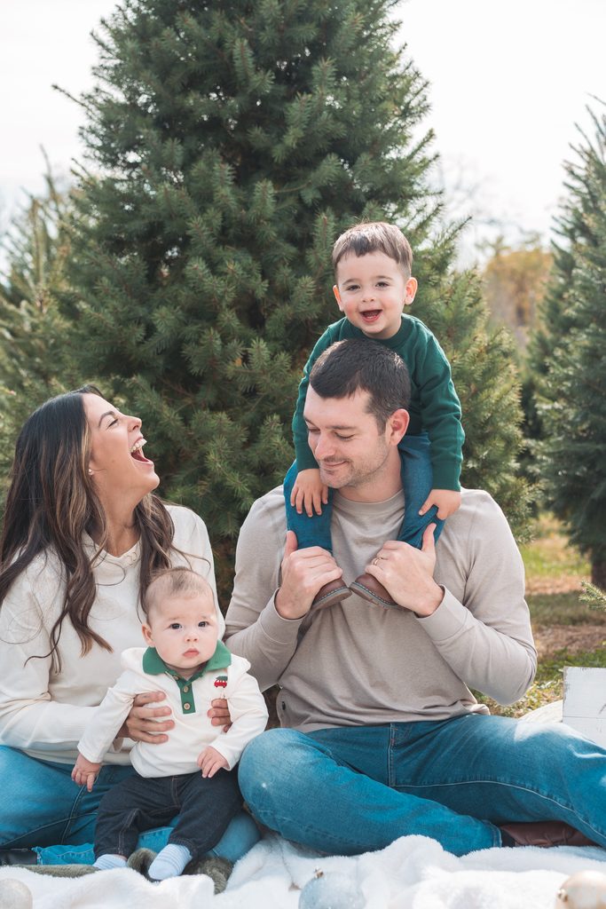 Family of four sitting together in front of an evergreen tree while their oldest son smiles from dad’s shoulders.