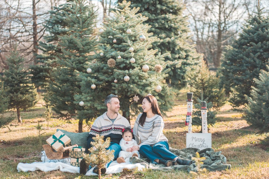 Family of three sitting beneath a decorated Christmas tree with wrapped gifts and festive holiday decor during a holiday mini session.