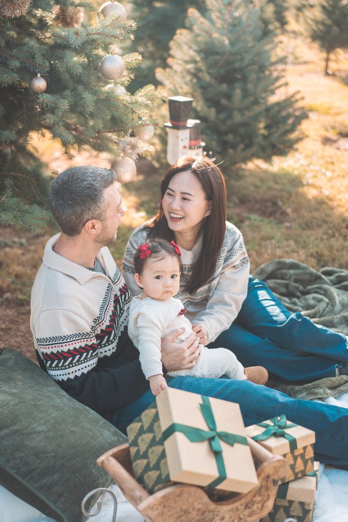 Family of three laughing together on a blanket surrounded by wrapped Christmas presents and holiday decorations.