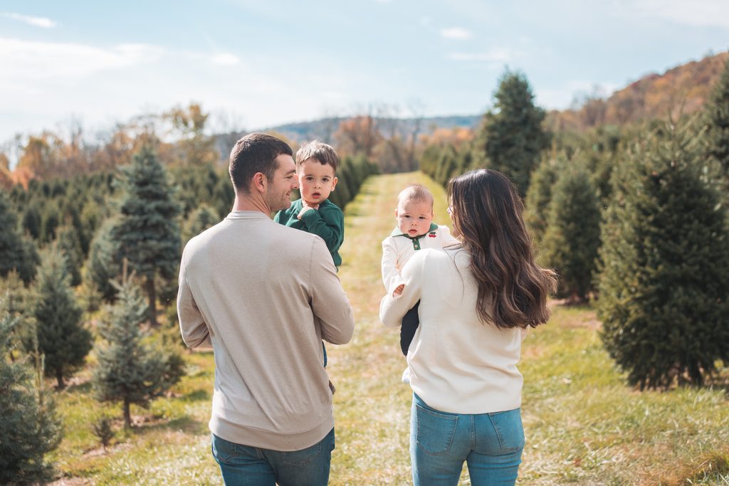 1. Family of four walking together between rows of Christmas trees while carrying their two young children.