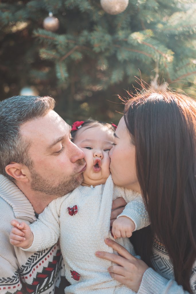 Mom and dad kissing their baby girl on both cheeks during a playful Christmas family photo.