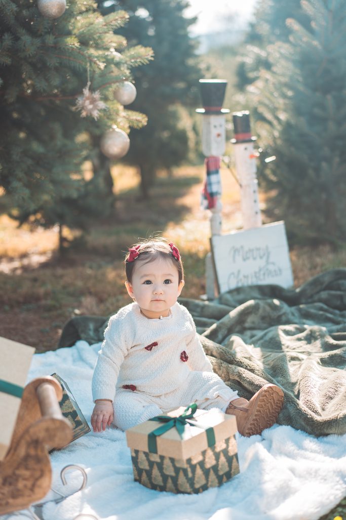 Baby girl with red bows sitting on a cozy blanket beside a wrapped gift during a festive holiday mini session.