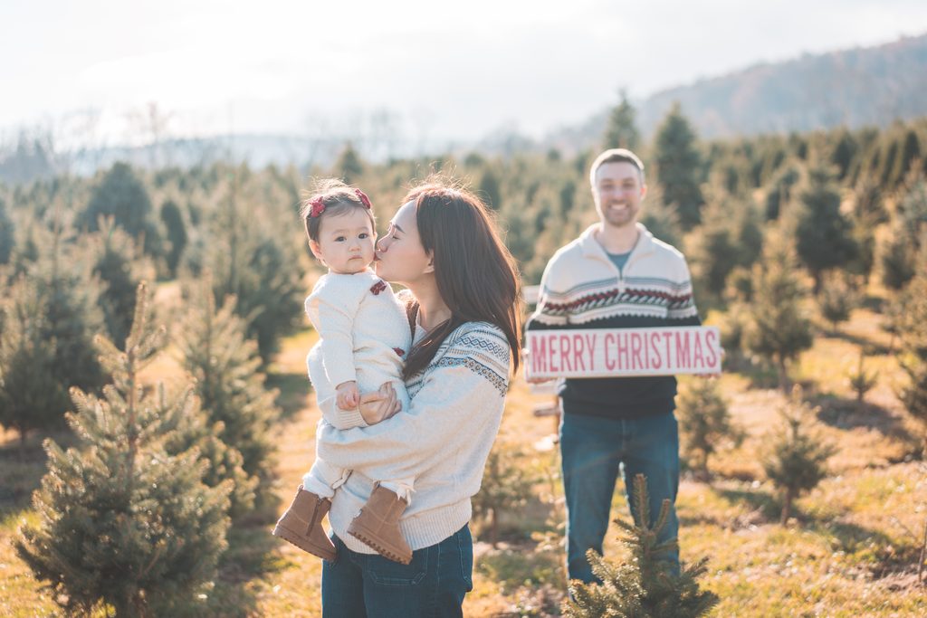 Mom kissing her baby while dad holds a Merry Christmas sign in the background during an outdoor holiday mini session.