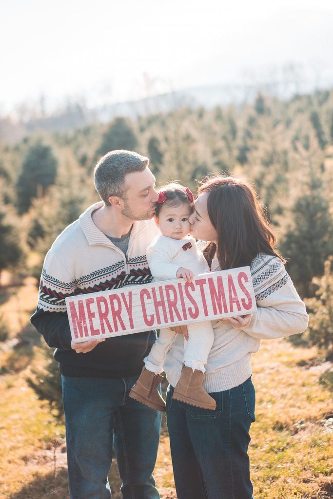 Parents kissing their baby girl while holding a Merry Christmas sign during a festive holiday mini session at a Christmas tree farm.