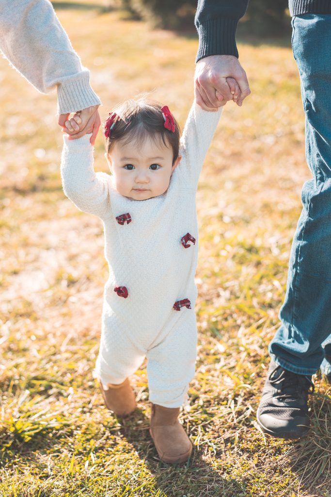 Baby girl in a white holiday outfit and red bows walking while holding both of her parents’ hands.