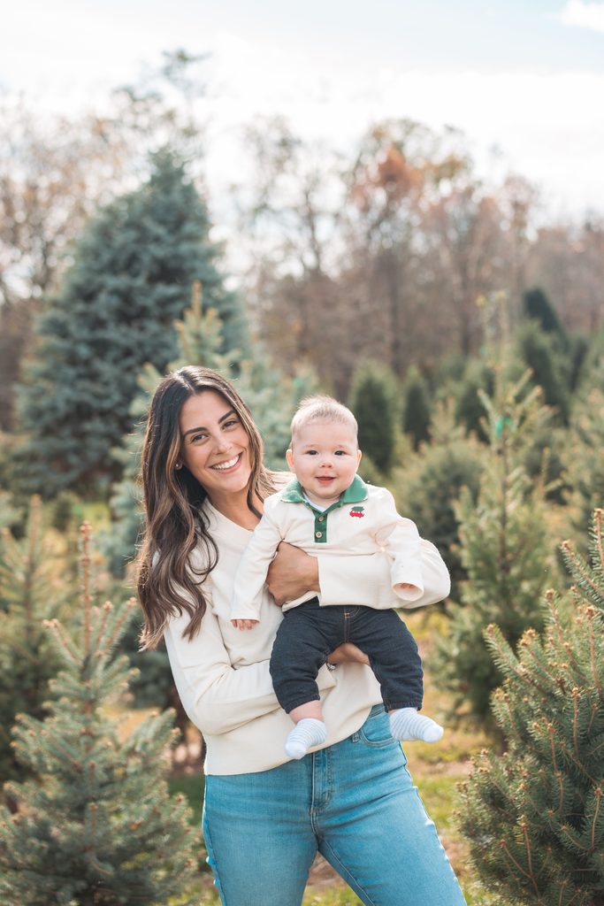 Mom smiling while holding her baby boy between evergreen trees during a holiday mini session at a Christmas tree farm.
