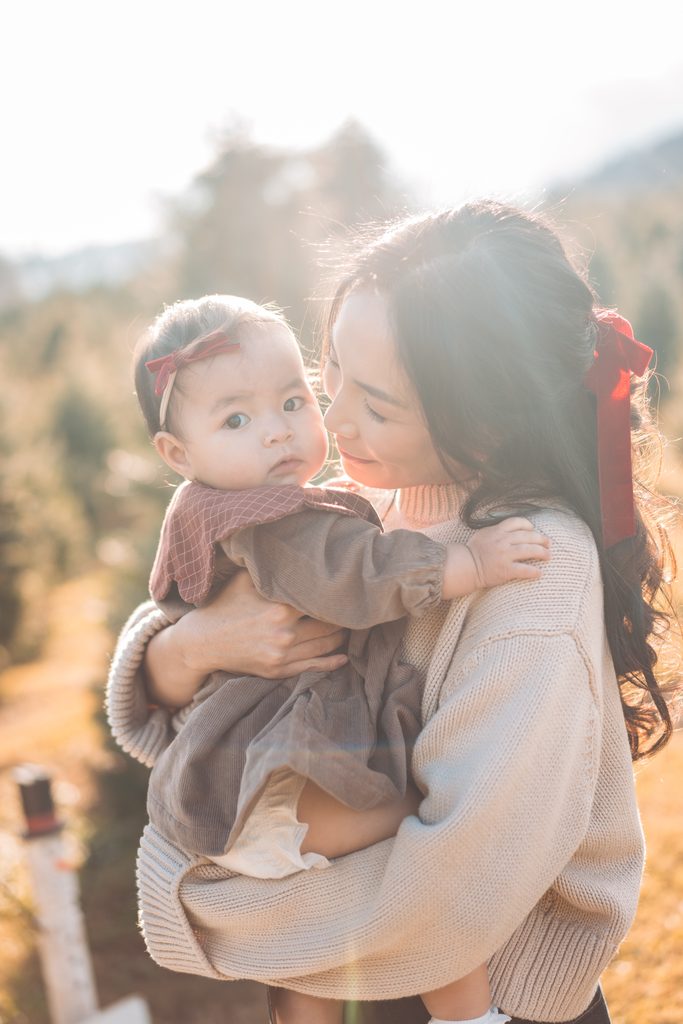 Mom holding her baby girl in warm sunlight during an outdoor holiday mini session at a Christmas tree farm.