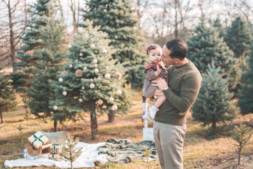 Dad holding and kissing his baby girl beside a decorated evergreen tree at a Christmas tree farm.