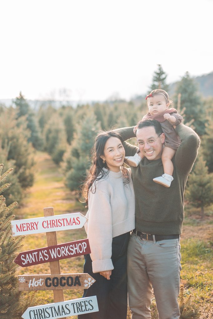 Parents smiling together with their baby girl on dad’s shoulders beside festive Christmas signs during a holiday mini session.