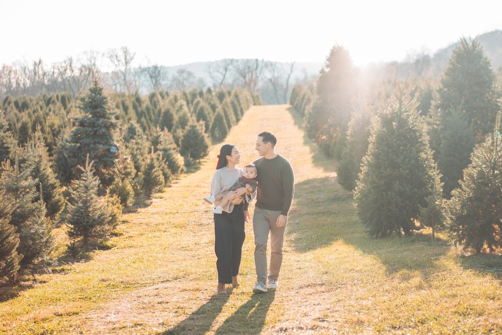 Mom and dad walking together through a Christmas tree farm while mom carries their baby.