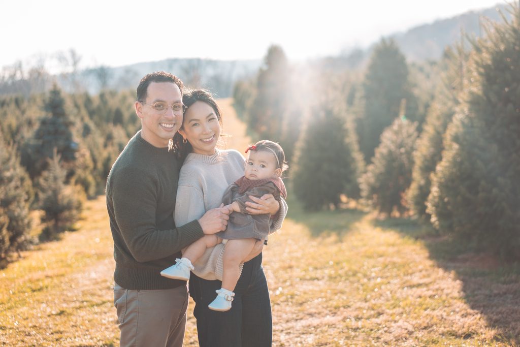Family of three smiling together while holding their baby girl between rows of Christmas trees during a sunny holiday mini session.